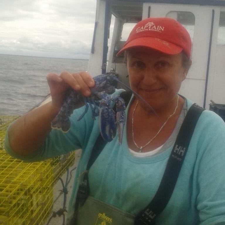 A smiling woman, wearing a red 'Captain' ball cap, stands on a boat holding a blue lobster.