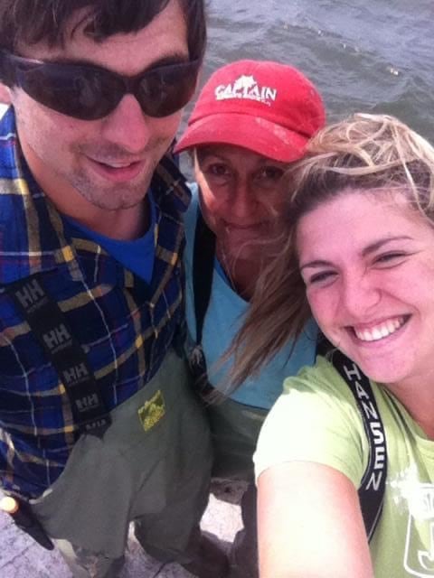 A selfie-style photo on a boat featuring a tall man in sunglasses and fishing waders, a woman wearing a 'Captain' ball cap, and a smiling young woman holding the camera with wind-blown hair.