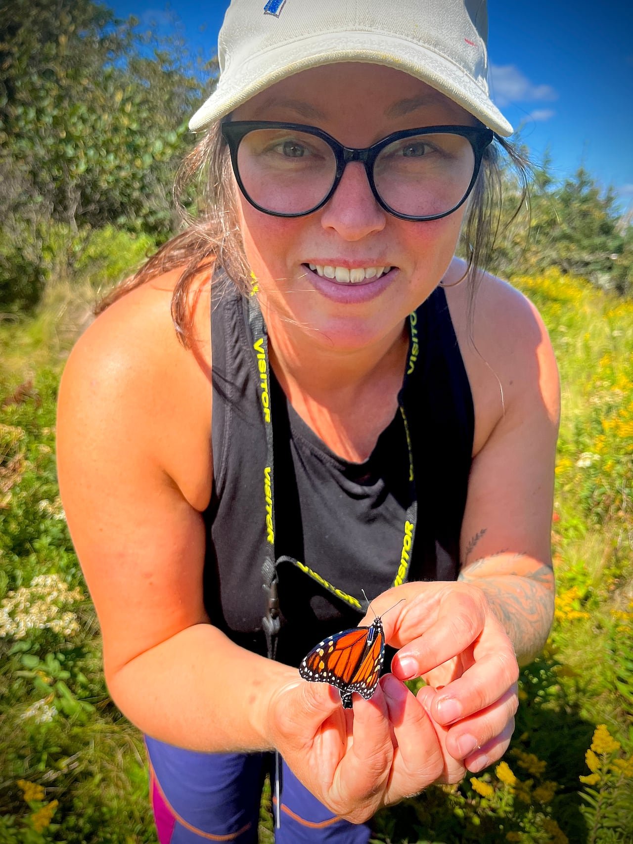 default-383 A woman with glasses and a white cap holding a monarch butterfly with a small tag on its thorax in a field of yellow wildflowers.