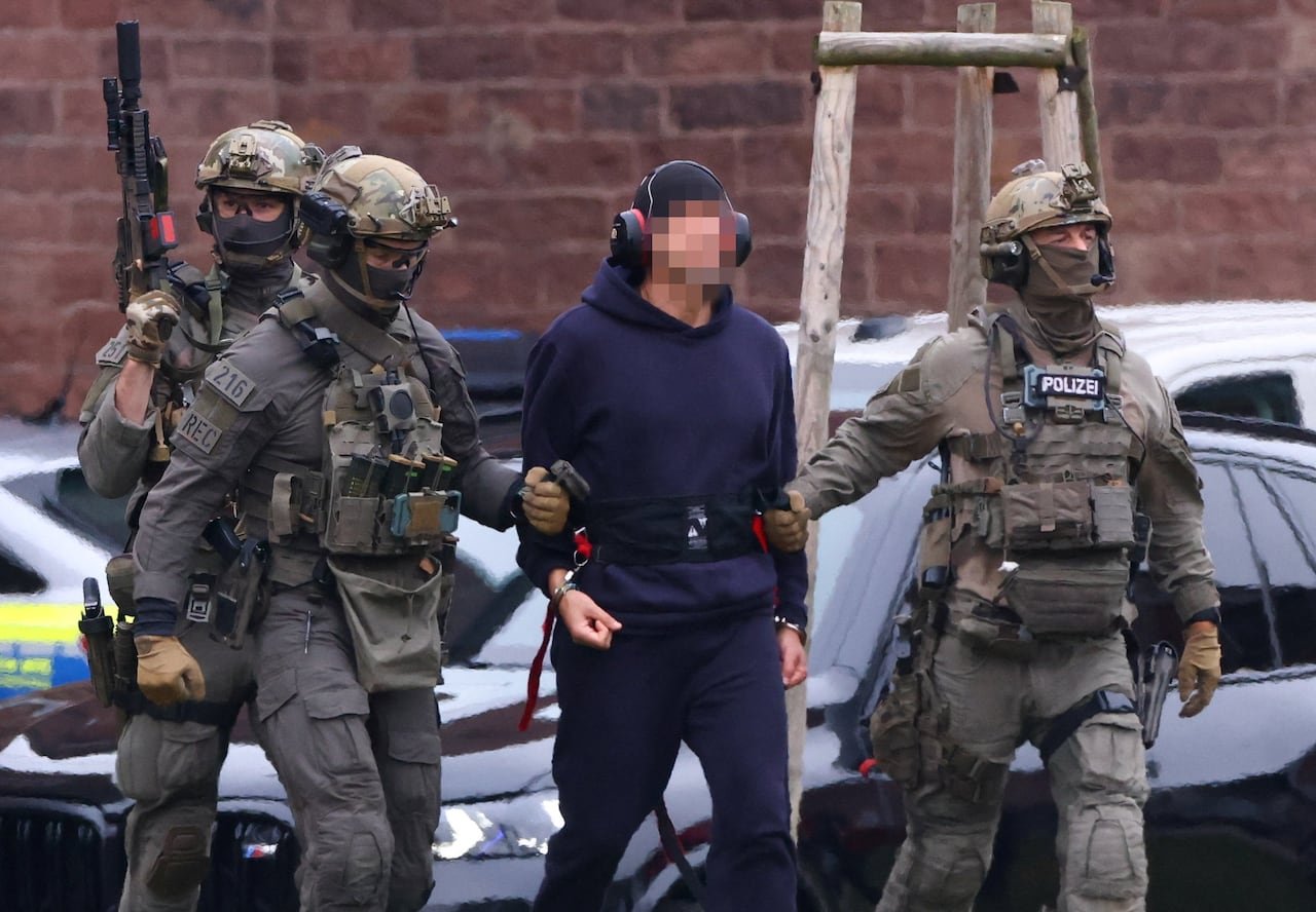 default-61 A man in a hoodie, ballcap, and headphones with blurred face escorted by three police officers in camouflage uniforms and holding guns.